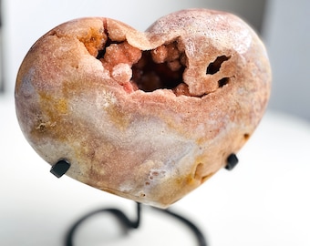 May include: A heart-shaped geode specimen featuring a smooth, polished exterior in shades of tan, beige, and soft brown, with a central cavity filled with small, textured, light orange and pink crystalline formations, mounted on a black metal stand.