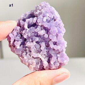 May include: A piece of light purple crystal with a bubbly texture, held in a hand. The crystal is composed of many small, round formations. The text "a1" is visible. The background is blurred, with hints of blue and white.