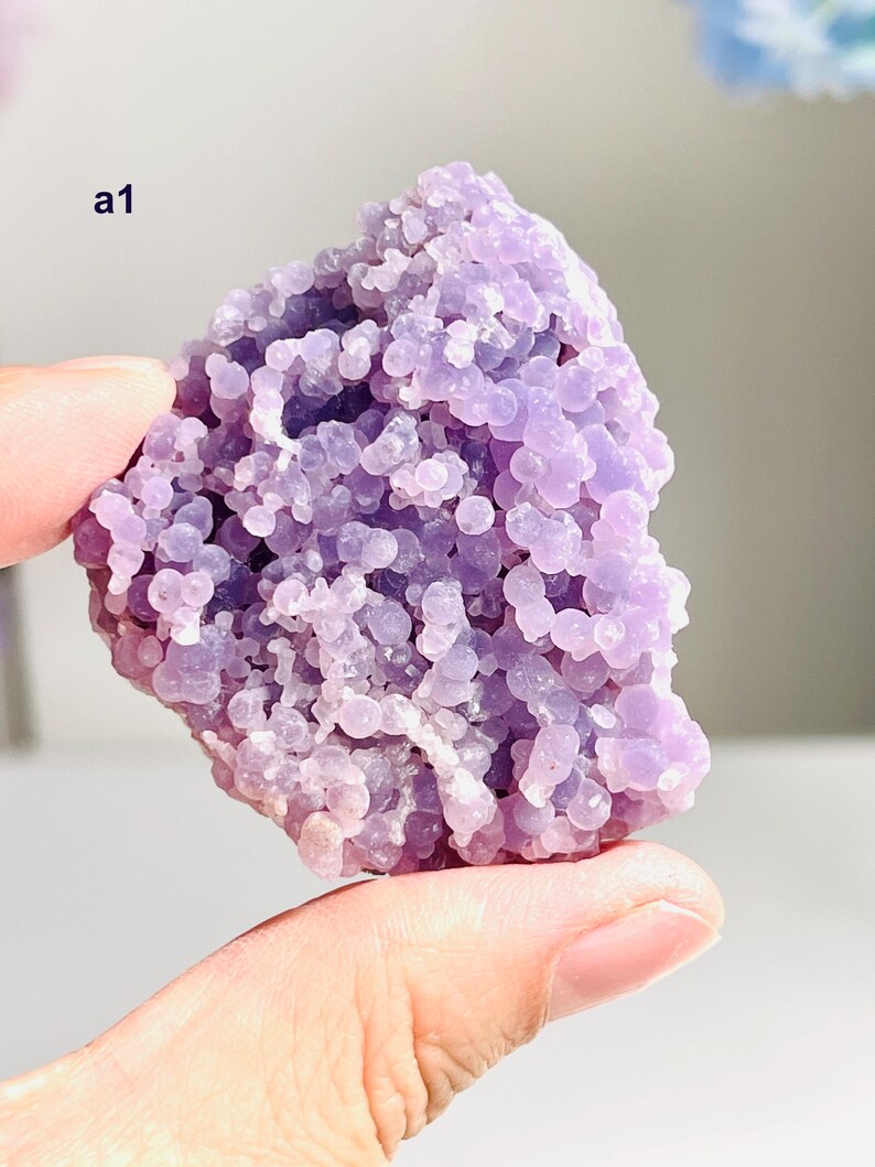 May include: A piece of light purple crystal with a bubbly texture, held in a hand. The crystal is composed of many small, round formations. The text "a1" is visible. The background is blurred, with hints of blue and white.