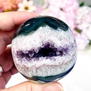 May include: A polished spherical stone featuring a deep green outer layer and a central horizontal band of jagged, light purple amethyst crystals. The sphere has a smooth, glossy surface finish that contrasts with the rough, crystalline interior texture.