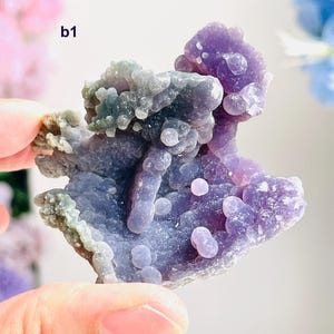 May include: A close-up of a purple and blue chalcedony geode. The geode has a bubbly, textured surface with areas of light green and lavender. The geode is held by a person's hand.