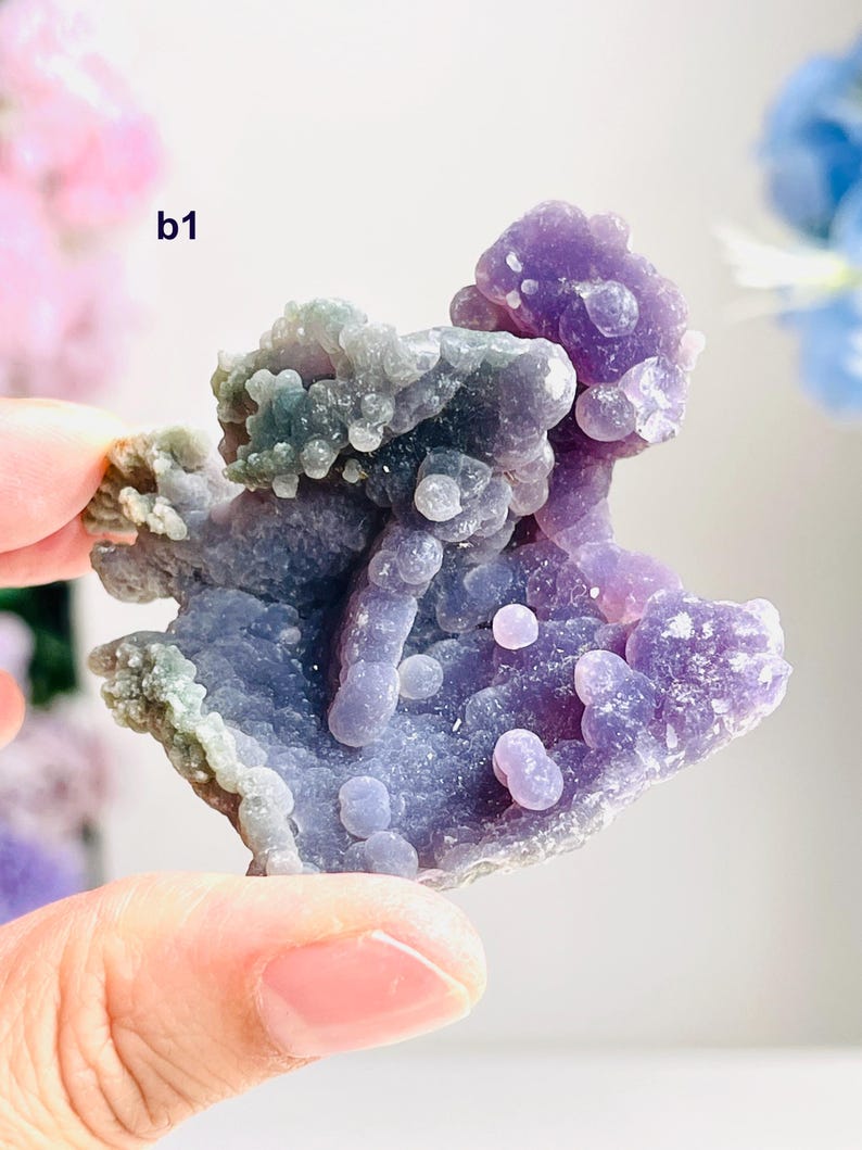 May include: A close-up of a purple and blue chalcedony geode. The geode has a bubbly, textured surface with areas of light green and lavender. The geode is held by a person's hand.