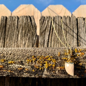 May include: A gold chain necklace with a pink and white gemstone pendant. The pendant is shaped like a triangle and has a smooth, polished surface. The necklace is hanging on a wooden fence.