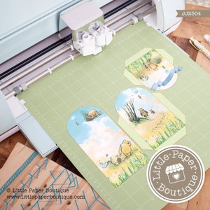 May include: A green cutting mat with three Winnie the Pooh themed paper die cuts. The die cuts are shaped like tags and feature Winnie the Pooh and his friends. The die cuts are being cut out by a Cricut Explore Air 2 machine. The machine is light blue and white. The text "Little Paper Boutique" is visible in the bottom right corner of the image.