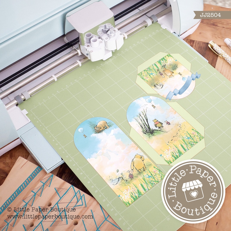 May include: A green cutting mat with three Winnie the Pooh themed paper die cuts. The die cuts are shaped like tags and feature Winnie the Pooh and his friends. The die cuts are being cut out by a Cricut Explore Air 2 machine. The machine is light blue and white. The text "Little Paper Boutique" is visible in the bottom right corner of the image.