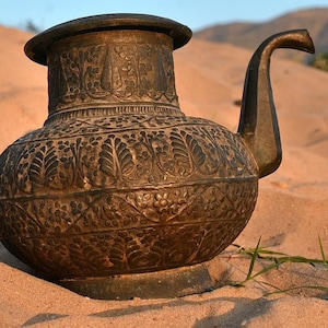 Puede incluir: Una olla de metal marrón oscuro grande y adornada con motivos, con un pico. La olla está sentada sobre una superficie arenosa.