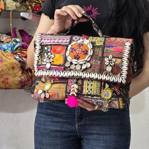 May include: A rectangular handbag featuring a patchwork of multicolored embroidered fabric, decorated with white cowrie shells, small circular mirrors, and silver-toned metal charms. A bright pink tassel hangs from the center of the front flap.