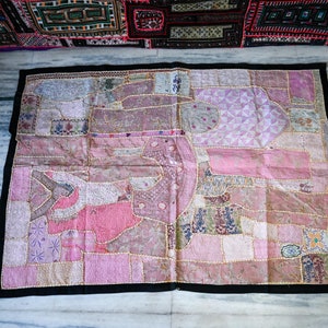 May include: A pink and white patchwork wall hanging with intricate embroidery details. The hanging is made of various fabrics and textures, creating a unique and colorful design.