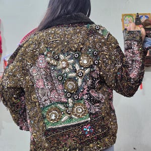 Può includere: Una giacca nera caratterizzata da ricami densi e complessi, fili metallici dorati e paillettes multicolori. Il pannello posteriore presenta un motivo floreale con grandi cristalli trasparenti sfaccettati, perline rosa e inserti in tessuto verde.