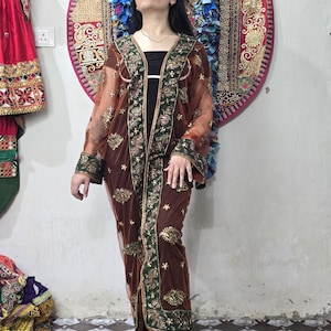 May include: A sheer brown duster coat featuring long sleeves and a dark green front placket. The garment is decorated with intricate gold embroidery and floral patterns throughout the fabric, extending to the cuffs and the wide vertical border.