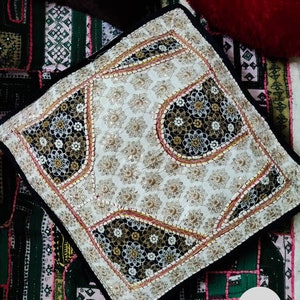 May include: A white and black patterned throw pillow with gold and silver embellishments. The pillow is on a colorful patchwork fabric.