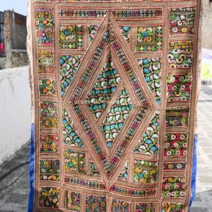 Op de afbeelding: Een kleurrijk patchwork-deken met ingewikkelde borduursels en spiegelversieringen. Het deken is gemaakt van verschillende stoffen in tinten roze, blauw, groen, geel en rood. Het deken wordt vastgehouden door de handen van een persoon.