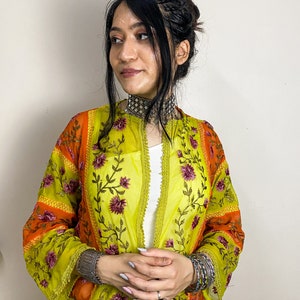 Puede incluir: Una chaqueta bordada verde, naranja y rosa con un patrón floral. La chaqueta tiene manga larga y un corte holgado. La modelo lleva la chaqueta sobre una camisa blanca.