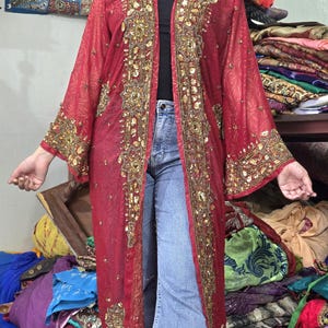 May include: A long, sheer, deep red open-front duster cardigan featuring intricate gold embroidery, sequins, and beadwork along the front placket and wide sleeves. The lightweight fabric has a subtle tonal pattern and falls to the mid-calf.