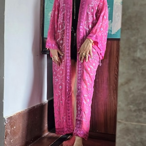 Könnte beinhalten: Ein langer, vorne offener Kimono-Duster in leuchtendem Pink mit aufwendigen silbernen Metallstickereien und Paillettenverzierungen. Das Kleidungsstück hat weite Dreiviertelärmel und eine gerade, fließende Silhouette bis zur Wadenmitte.