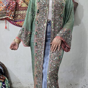 May include: A long, open-front sheer duster in a sage green and dusty pink gradient, featuring intricate silver sequin embroidery, metallic thread detailing, and circular green accents along the front placket and wide, flowing sleeves.