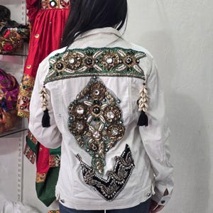 May include: A white denim jacket featuring elaborate back embroidery with green, gold, and black beads, sequins, and clear crystals. The design includes a horizontal band across the shoulders, a large central diamond-shaped motif, and two hanging shell tassels.