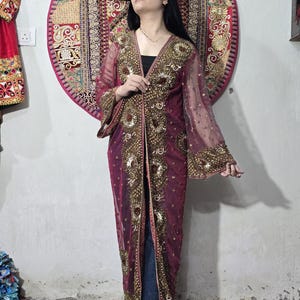 May include: A long, sheer burgundy duster featuring intricate gold embroidery, sequins, and beadwork along the front panels and wide sleeves. The garment is open-front, floor-length, and crafted from a lightweight, translucent fabric with a metallic finish.