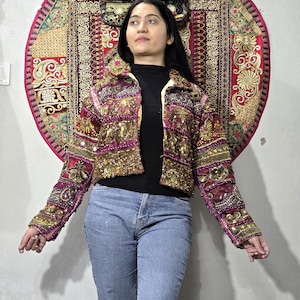 May include: A cropped jacket featuring intricate embroidery, gold sequins, and colourful fabric patches in shades of magenta, gold, and deep red. The garment has long sleeves, a collared neckline, and a button-down front, showcasing detailed needlework.