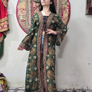 May include: A long, sheer, dark green open-front duster coat featuring gold floral embroidery and intricate mirror work along the edges and sleeves. The garment has wide, flowing sleeves and a straight, floor-length silhouette.