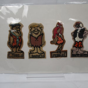 May include: Four vintage felt patches featuring the characters from the cartoon "The Flintstones". The patches are brown and feature Fred, Barney, Pebbles, and Bamm-Bamm. The patches are labeled with the character's names.