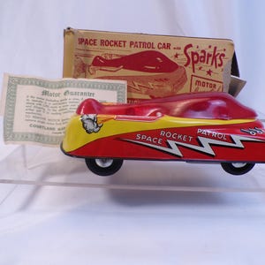 May include: A red and yellow toy car with a "Space Rocket Patrol" design and a "Sparks Motor" label on the box. The car has a wind-up motor and is made of tin.