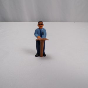 May include: A blue figurine of a person standing behind a brown podium. The person is wearing a blue shirt, a dark tie, and dark pants. The person has brown hair and is wearing glasses.