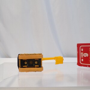 May include: A yellow and black toy bulldozer with a textured track design. The bulldozer has a yellow arm and blade. A red and white box with Japanese text is in the background.