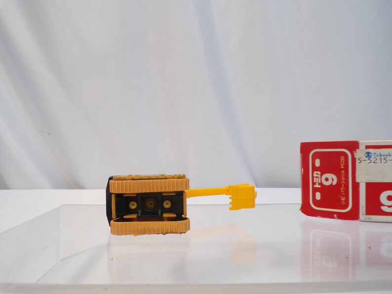 May include: A yellow and black toy bulldozer with a textured track design. The bulldozer has a yellow arm and blade. A red and white box with Japanese text is in the background.