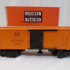 May include: Orange Lionel electric train boxcar with "AT&SF 63132" and "Santa Fe" logos. The boxcar has a black door and a ladder on the side. The boxcar is in a box with "Lionel Electric Trains 3464" printed on it.