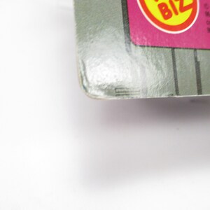 May include: Close-up of a product package corner. The package has a gray and pink color scheme with a yellow and red circular logo. Vertical gray lines are visible on the package. The text "BIZ" is visible in the logo.