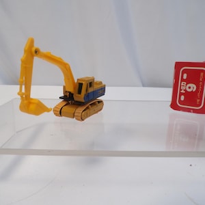 May include: A yellow and blue toy excavator with a movable arm and bucket. The toy is on a clear surface, with a red and white box with Japanese text in the background. The excavator is a miniature construction vehicle.