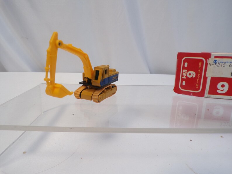 May include: A yellow and blue toy excavator with a movable arm and bucket. The toy is on a clear surface, with a red and white box with Japanese text in the background. The excavator is a miniature construction vehicle.