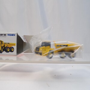 May include: A yellow and black Komatsu articulated dump truck toy, partially visible within a clear plastic wrapper. The toy truck is next to its box, which features a picture of the truck and Japanese text.