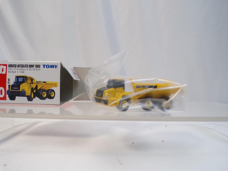 May include: A yellow and black Komatsu articulated dump truck toy, partially visible within a clear plastic wrapper. The toy truck is next to its box, which features a picture of the truck and Japanese text.