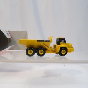 May include: A yellow toy dump truck with black wheels and a black cab. The truck has the number "400" printed on the side. The toy is sitting on a clear surface, with a partially visible box in the background.