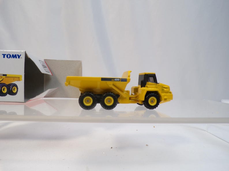 May include: A yellow toy dump truck with black wheels and a black cab. The truck has the number "400" printed on the side. The toy is sitting on a clear surface, with a partially visible box in the background.