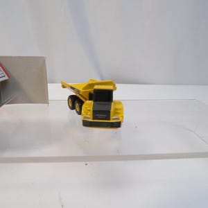 May include: A yellow and black toy dump truck with black wheels. The toy truck is on a clear surface. A white and red box is in the background.