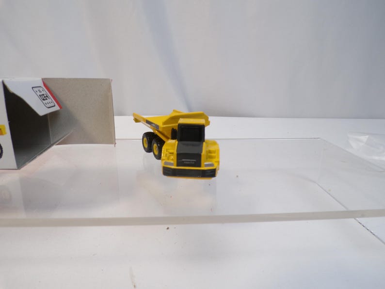 May include: A yellow and black toy dump truck with black wheels. The toy truck is on a clear surface. A white and red box is in the background.