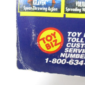 Pu&ograve; includere: Primo piano di un logo Toy Biz blu e giallo con il numero di telefono 1-800-634-75 stampato sotto. Il logo &egrave; un cerchio con le parole "Toy Biz" in lettere bianche.