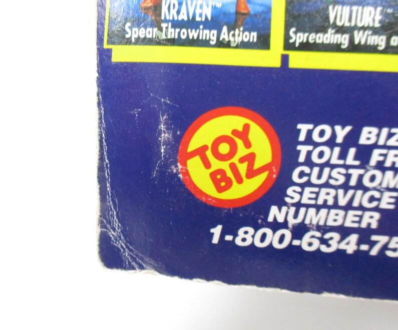 Pu&ograve; includere: Primo piano di un logo Toy Biz blu e giallo con il numero di telefono 1-800-634-75 stampato sotto. Il logo &egrave; un cerchio con le parole "Toy Biz" in lettere bianche.