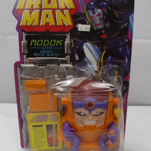 May include: A plastic action figure of MODOK, a Marvel Comics supervillain, in a seated position. MODOK is wearing a purple and orange suit with a large head and a red gem on his forehead. The figure is packaged in a clear plastic blister pack with a cardboard backing. The packaging features the Marvel Comics logo and the text "Iron Man" and "MODOK with Energy Brain Blasts".