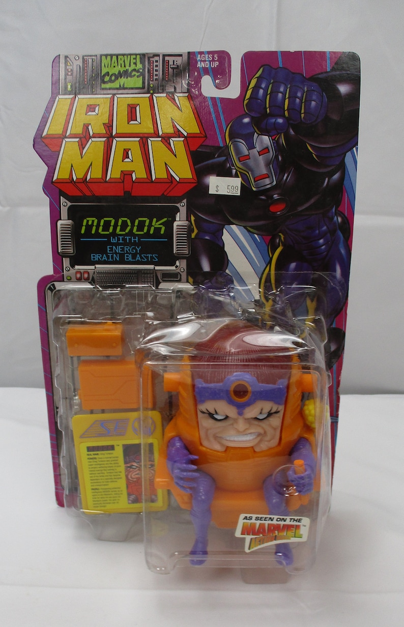May include: A plastic action figure of MODOK, a Marvel Comics supervillain, in a seated position. MODOK is wearing a purple and orange suit with a large head and a red gem on his forehead. The figure is packaged in a clear plastic blister pack with a cardboard backing. The packaging features the Marvel Comics logo and the text "Iron Man" and "MODOK with Energy Brain Blasts".