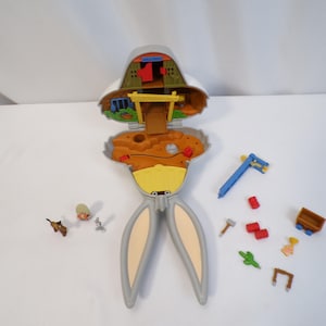 May include: A Bugs Bunny-themed toy playset, featuring a gray and yellow rabbit-shaped case that opens to reveal a miniature interior scene. Includes small accessories like a pickaxe, cart, and figures. The playset is designed to resemble a cartoon setting.
