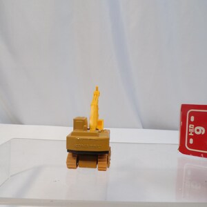May include: A small, tan and yellow toy excavator with black treads and a raised arm. The toy is on a clear surface with a red and white box in the background. The excavator has the word "KOMATSU" printed on it.