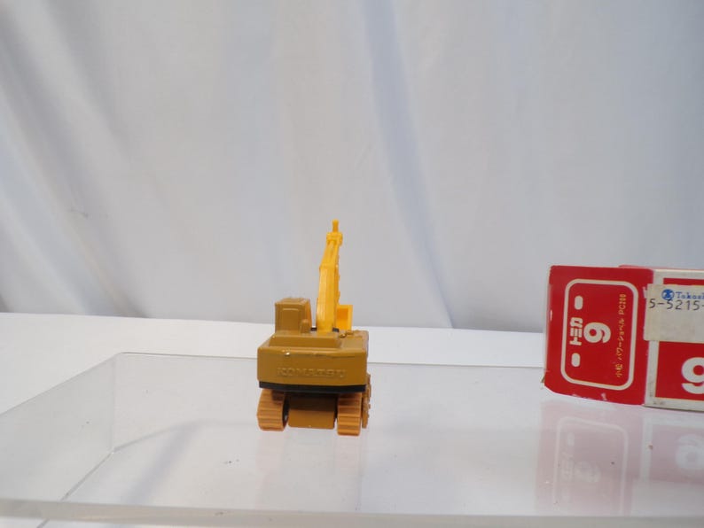 May include: A small, tan and yellow toy excavator with black treads and a raised arm. The toy is on a clear surface with a red and white box in the background. The excavator has the word "KOMATSU" printed on it.