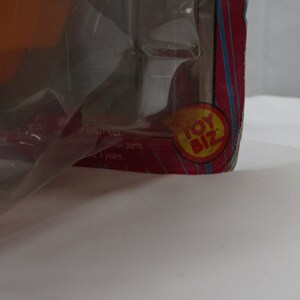 May include: Close-up of a red and yellow Toy Biz logo on a pink and blue striped background. The logo is in a circle with the words "Toy Biz" in white letters.