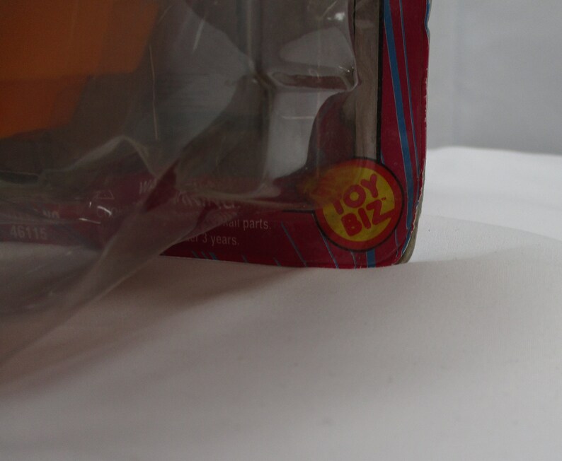 May include: Close-up of a red and yellow Toy Biz logo on a pink and blue striped background. The logo is in a circle with the words "Toy Biz" in white letters.