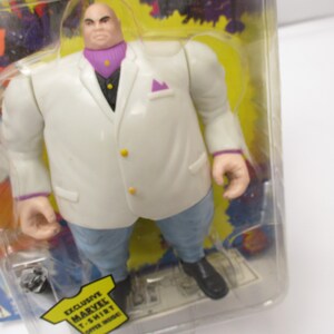 Pu&ograve; includere: Una action figure bianca di Kingpin dei Marvel Comics. La figura indossa un abito bianco con una camicia e una cravatta viola. La figura &egrave; in piedi su uno sfondo giallo con le parole "Exclusive Marvel T-Shirt" stampate in basso.
