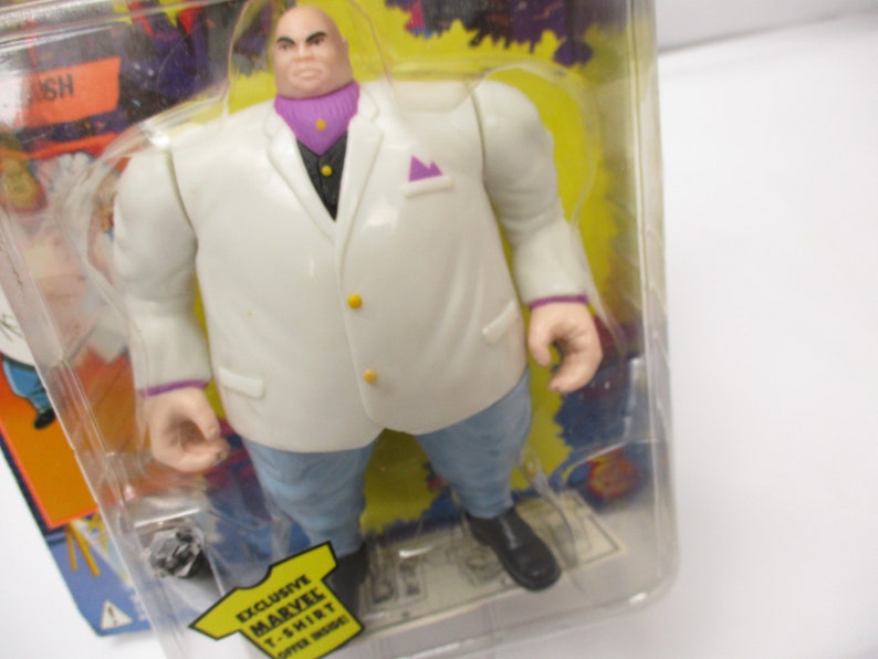 Pu&ograve; includere: Una action figure bianca di Kingpin dei Marvel Comics. La figura indossa un abito bianco con una camicia e una cravatta viola. La figura &egrave; in piedi su uno sfondo giallo con le parole "Exclusive Marvel T-Shirt" stampate in basso.